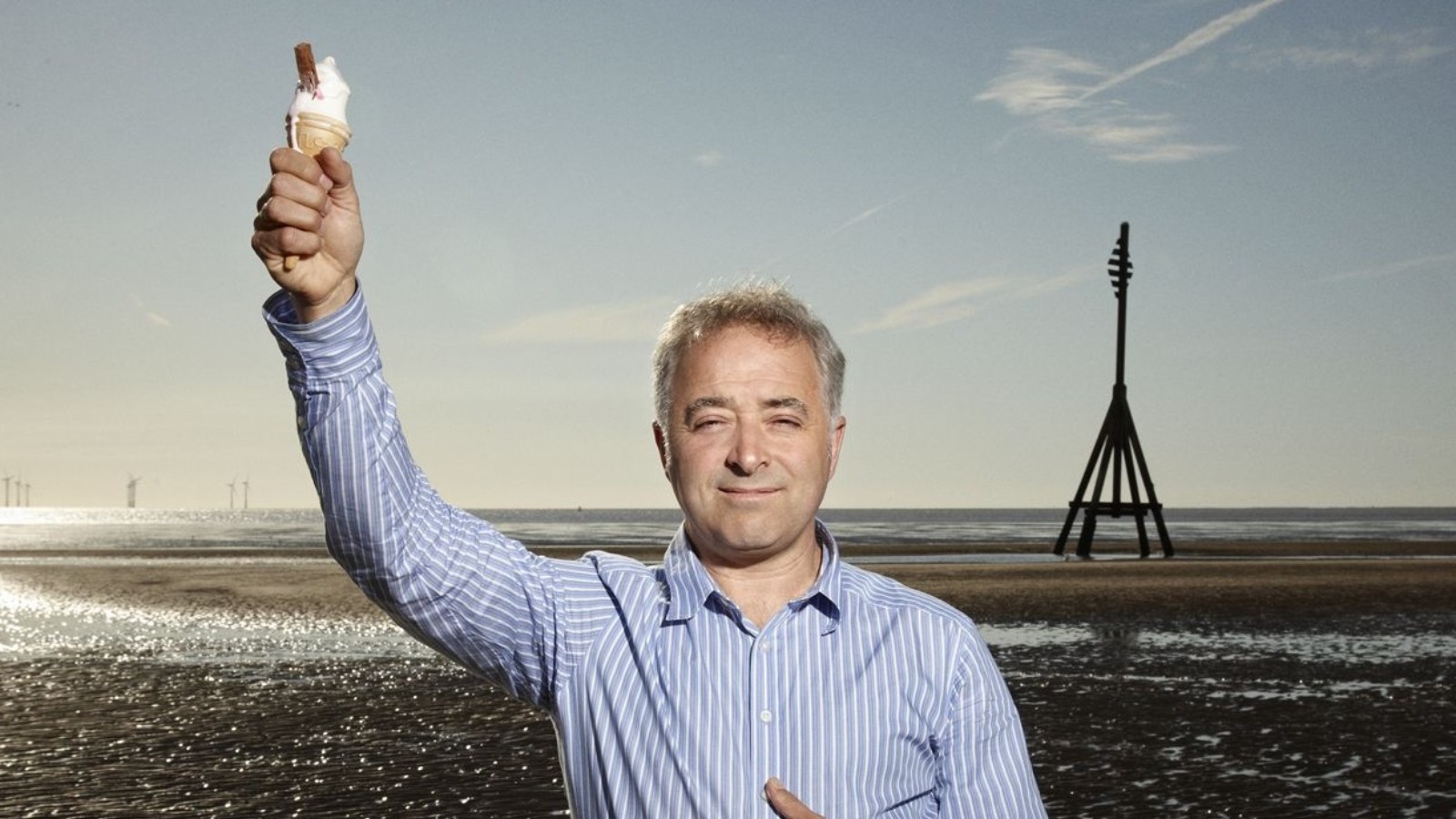 Screenwriter and Novelist Frank Cottrell Boyce stands on a beach against a blue sky holding an ice cream cone with a flake in the air.