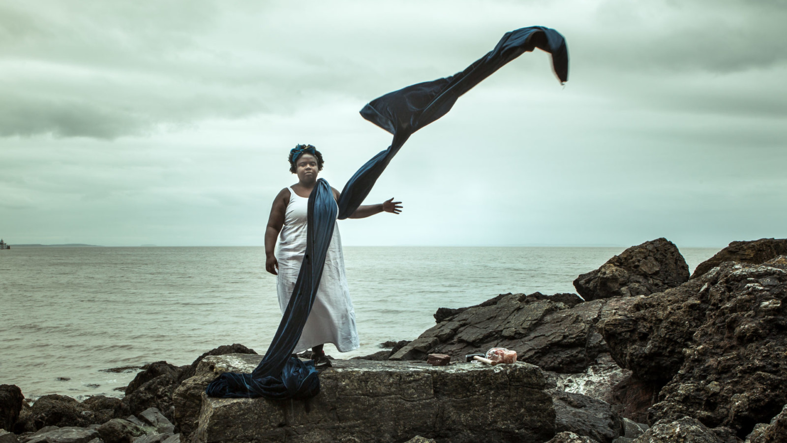 Artist Selina Thompson stands on rocky ground next to a grey sea and clouded sky. She is draped in a long piece of navy fabric which bellows and flies in the wind. Next to Selina on the ground is a mallet and a large chunk of pink salt.
