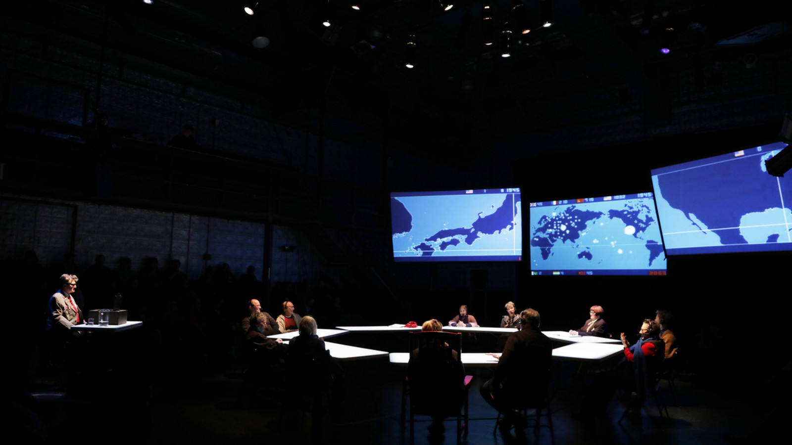 In a dark theatre 3 large screens suspended to one side. The screen show blueprint maps. A large circle of tables has a person sat at each table and are lit from above. To the side of the stage another table with one person faces towards the circle.