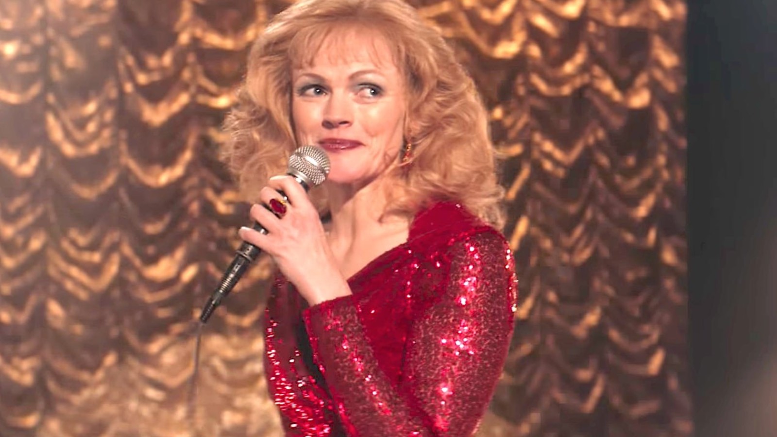A woman with blonde hair wears a red glittering dress and holds a microphone smiling. She stands in front of a gold, scalloped curtain.