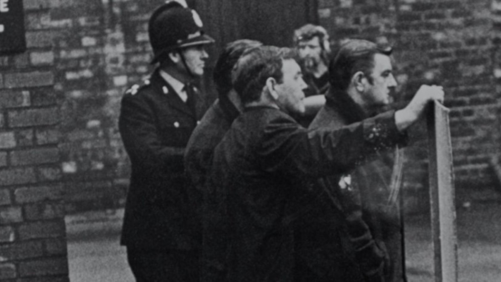 Still image from the film Rank and File. A black and white image of a group of men leaning on a fence and behind them a police officer.