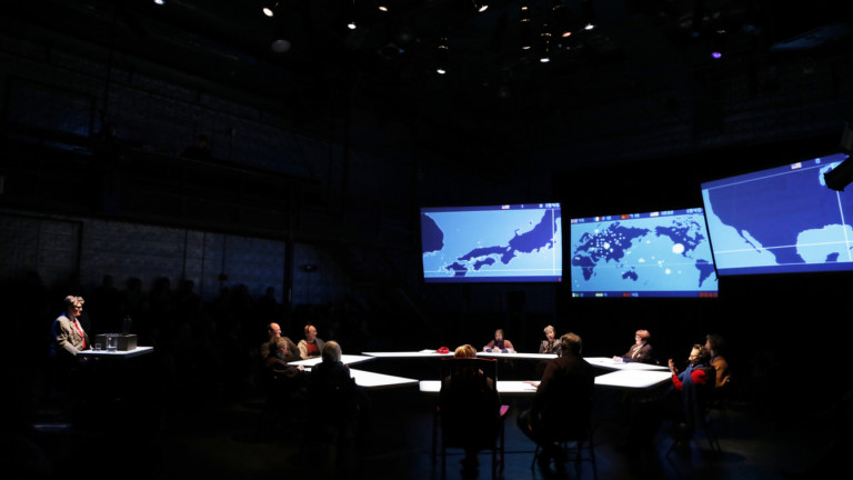 In a dark theatre 3 large screens suspended to one side. The screen show blueprint maps. A large circle of tables has a person sat at each table and are lit from above. To the side of the stage another table with one person faces towards the circle.