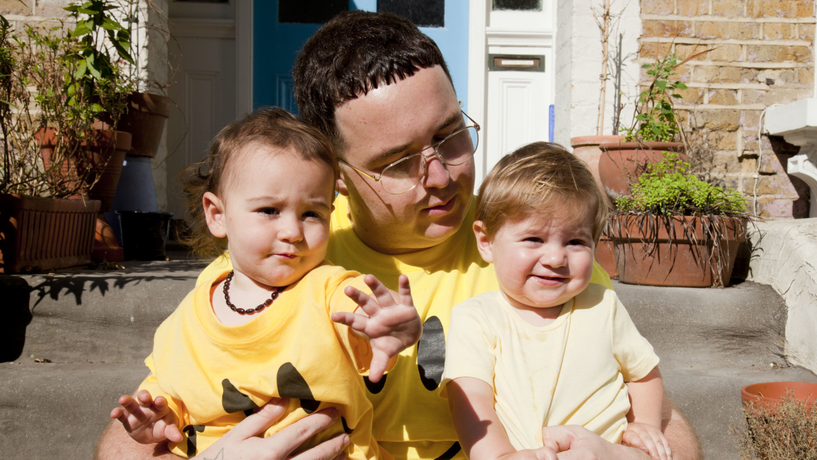 A person with short dark hair and glasses sits on stone steps in front of a blue residential doorway. They are holding two small children with brown hair, one in each arm. They all wear yellow t-shirts. One of the children and the adult wear a yellow t-shirt with a black smiley face on. The adult is looking down at one of the children as the other child reaches out towards the camera.