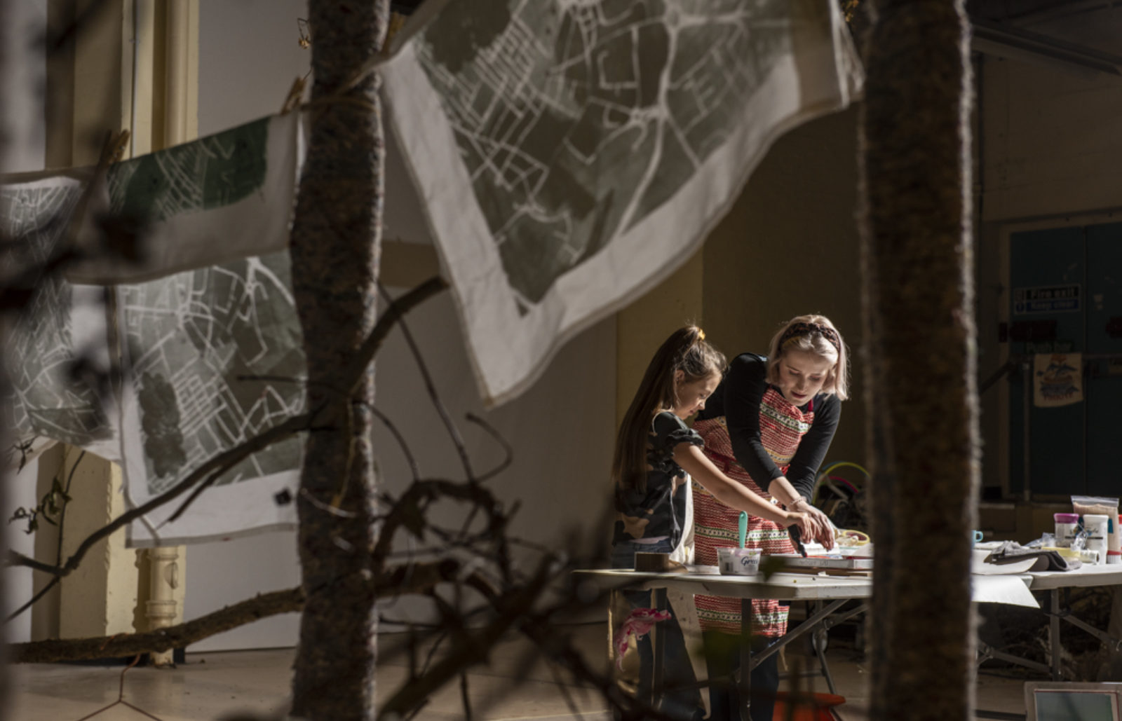 Two people, a young person and an adult stand in the center of a room screen printing. Around them hang some pieces of cloth with a map design printed on them.