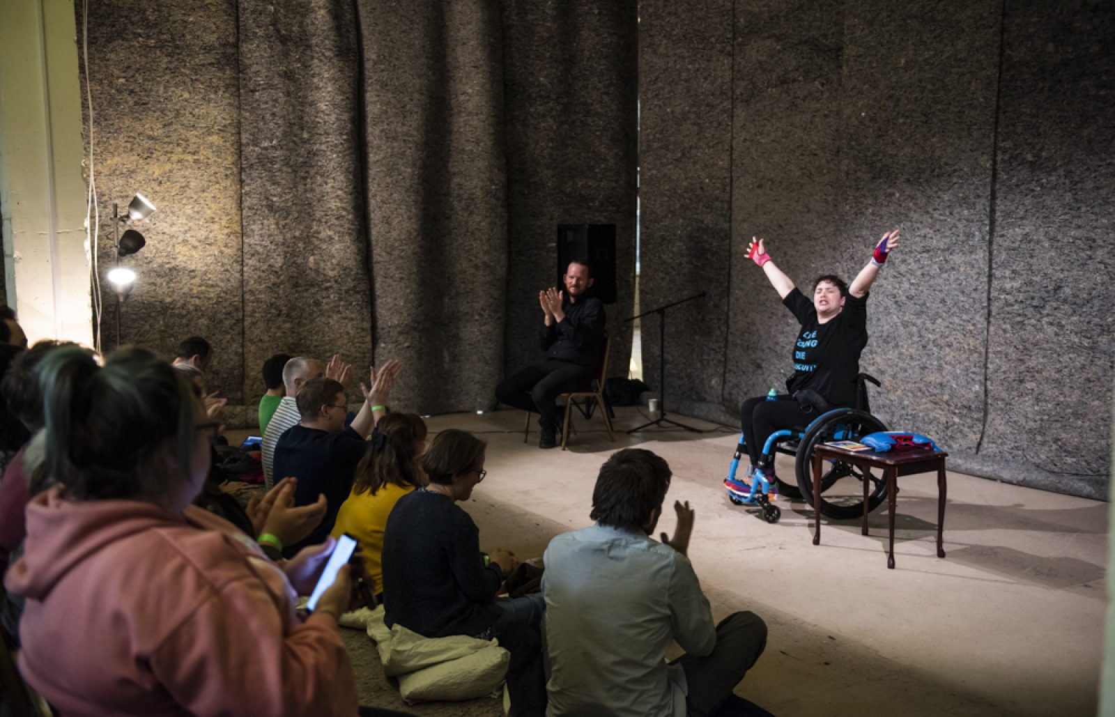 A person in a manual wheelchair is on stage with their hands in the air. An audience of people watch and clap.