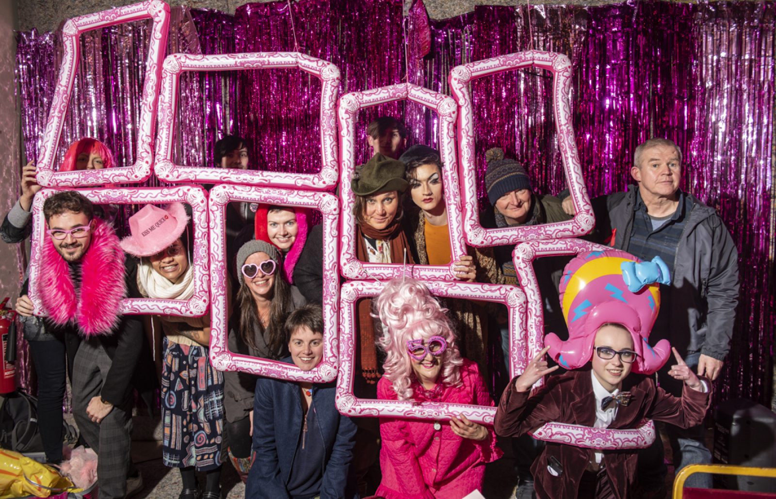 In front of a bright pink glitter curtain a group of people stand and kneel with pink accessories on and framed with pink inflatable frames. They are smiling and posing for a photograph.