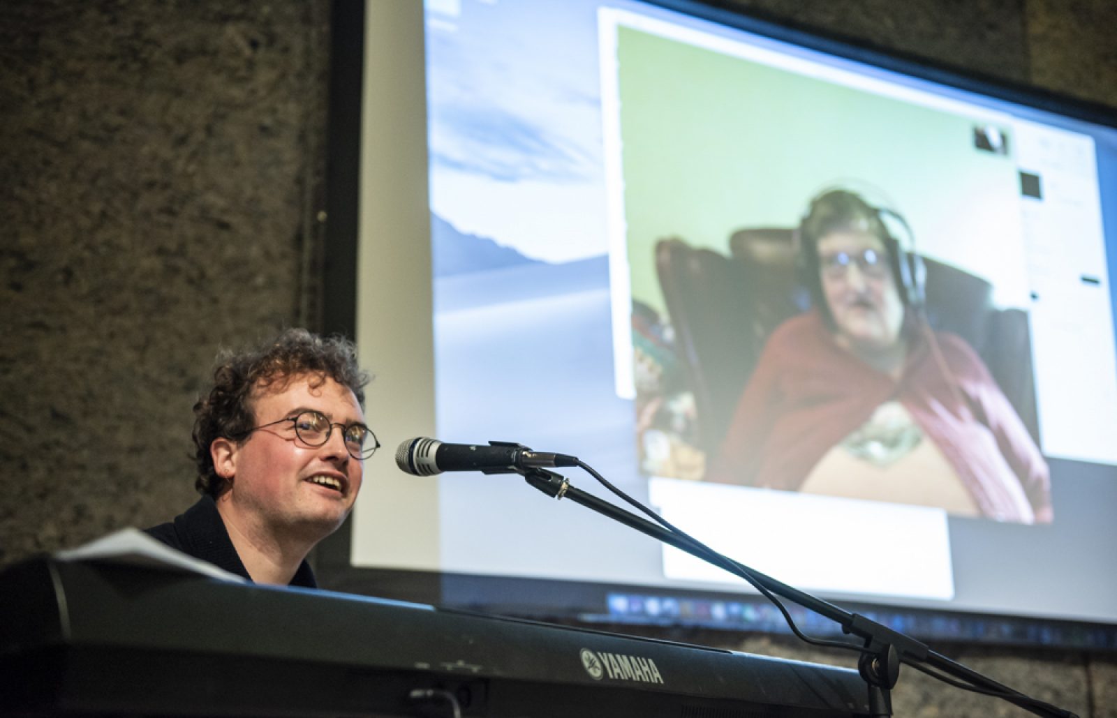 A person with short curly hair and glasses sits behind a Yamaha keyboard and a microphone smiling. On a projector screen behind them there is a person sitting in an arm chair with headphones on.
