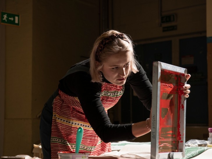 A person with blonde hair in a bob haircut, wearing a headband, a black long sleeved top and a multi-coloured apron leans down to look at a screen printing frame. This is a wooden frame with canvas painted red in the centre.