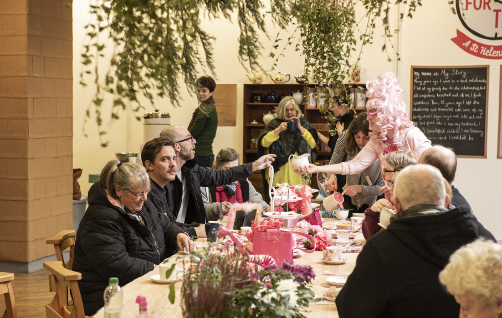 A long table set with a pink table cloth, tea set and decorations. A number of people sit around the table. A woman in a pink wig is serving tea. Greenery hangs down and there is lavender on the table.