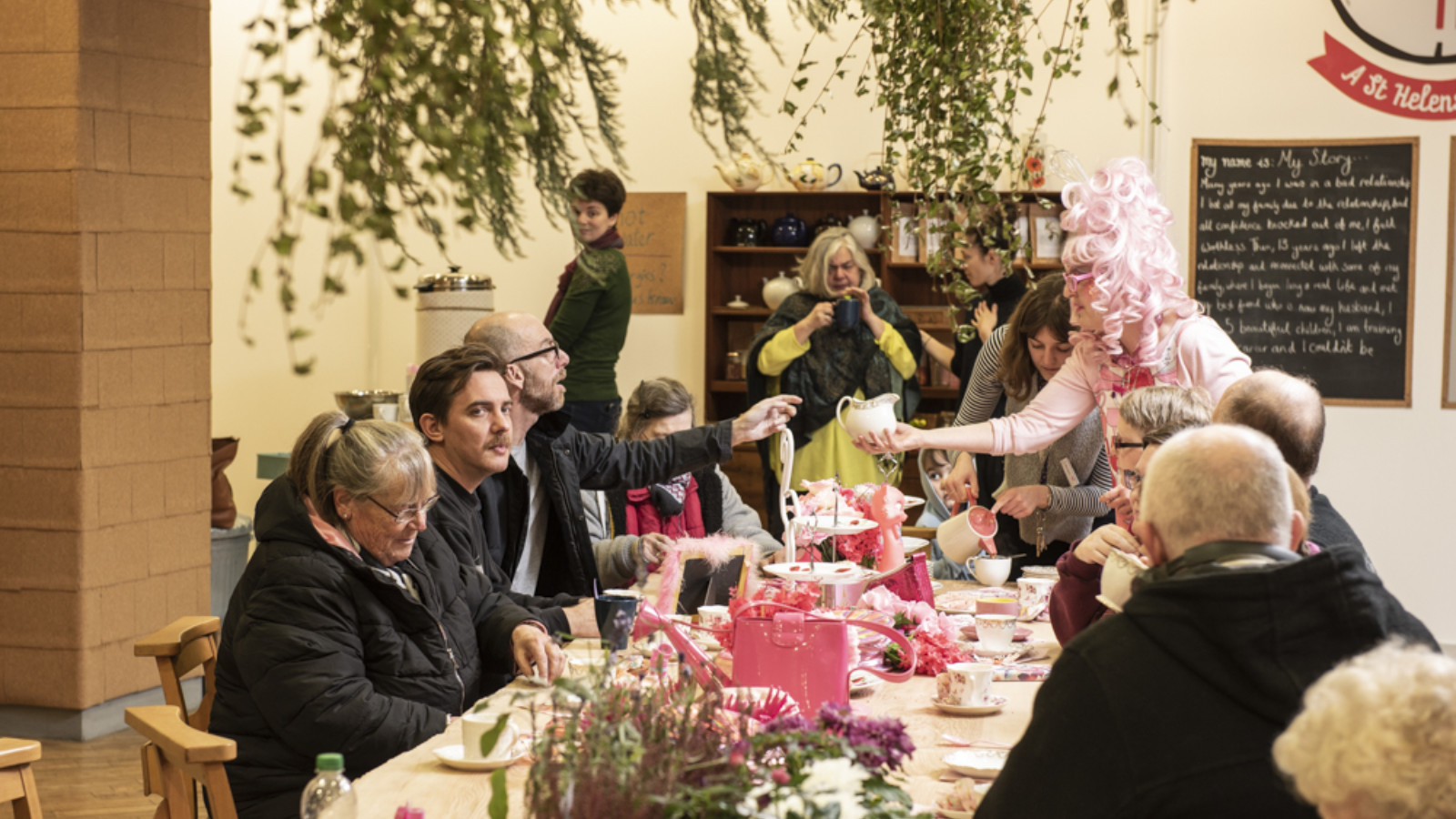 A long table set with a pink table cloth, tea set and decorations. A number of people sit around the table. A woman in a pink wig is serving tea. Greenery hangs down and there is lavender on the table.