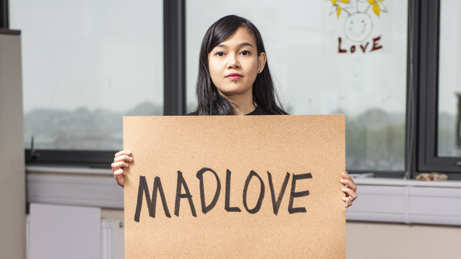Indonesian artist Hana Madness stands facing the camera holding a cork placard that reads MADLOVE.|