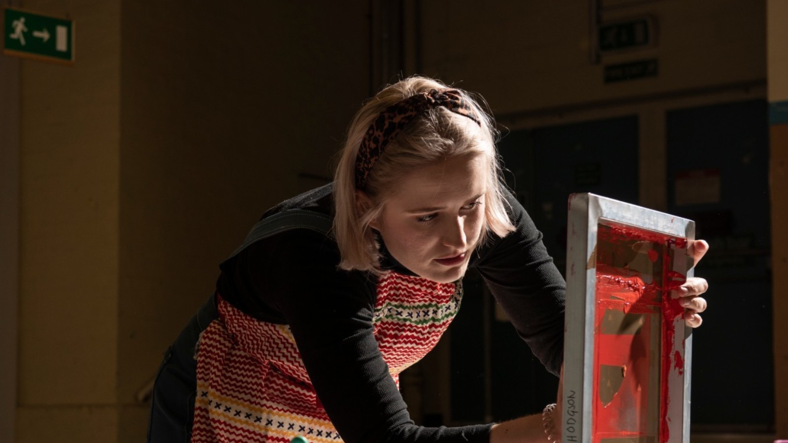 A person with blonde hair in a bob haircut, wearing a headband, a black long sleeved top and a multi-coloured apron leans down to look at a screen printing frame. This is a wooden frame with canvas painted red in the centre.