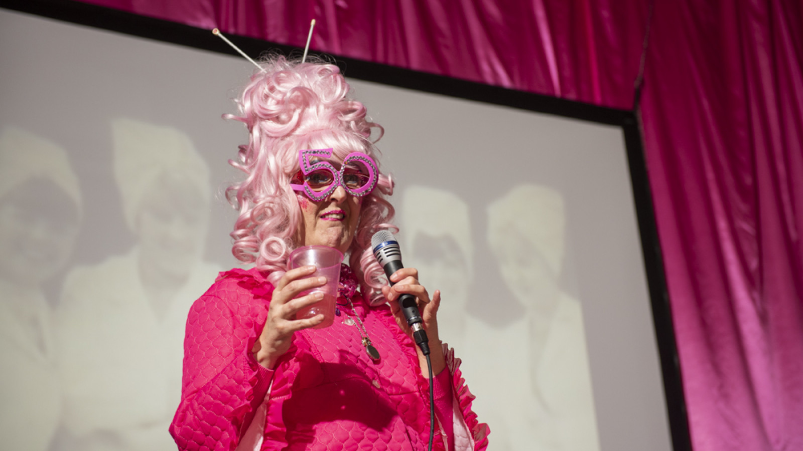 A middle aged white woman stands on a stage with a mic in her right hand and a pink drinking glass in her left. She is dressed head-to-toe in bright pink; her bee-hive hair do is pink; there are a pair of knitting needles stuck in the top of her high hair do, these are pink too; she also wears a comedy pair of glasses whose oversized frames form the numbers 50 in pink