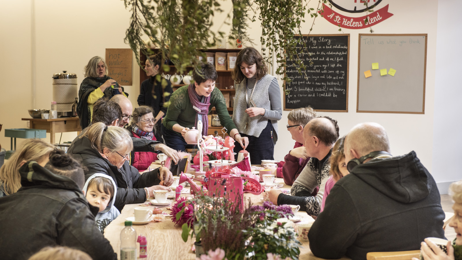 People sit around a long table in a light filled room. The people are of all ages from a young child to older people. The table is covered in flowers and everyone has a cup and saucer in front of them. Two women stand at the end of the table serving cups of tea and are smiling.