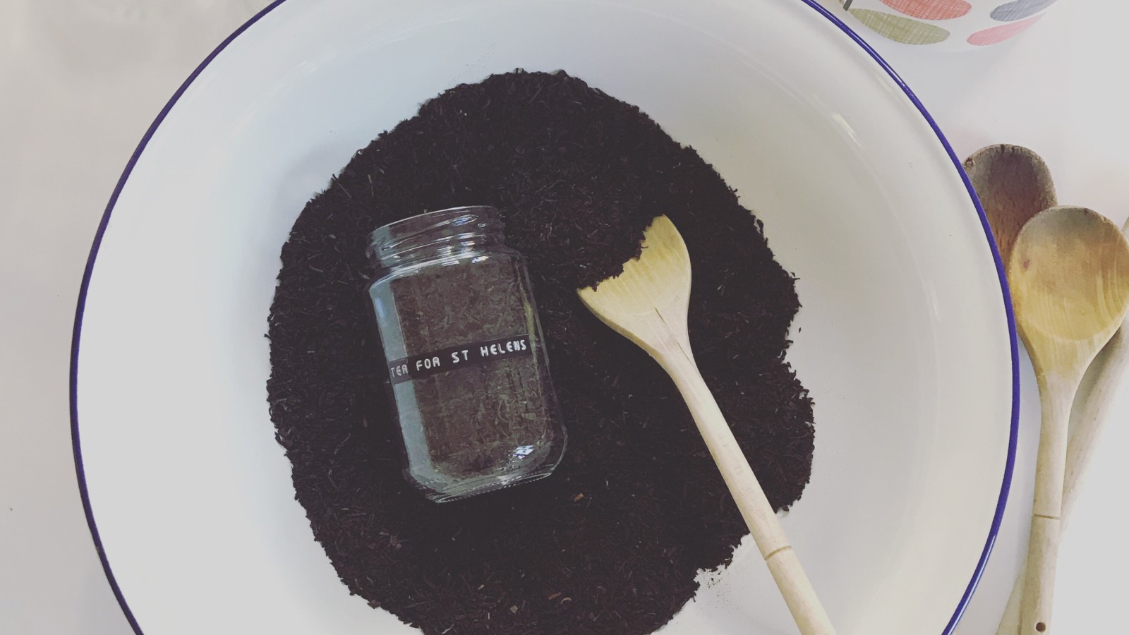 A glass jar with a label on it reading 'Tea for St Helens' lies on tea leaves at the bottom of a large mixing bowl with a blue rim. There is also a wooden spoon next to the jar in the tea leaves and other glass jars and spoons surrounding the bowl.