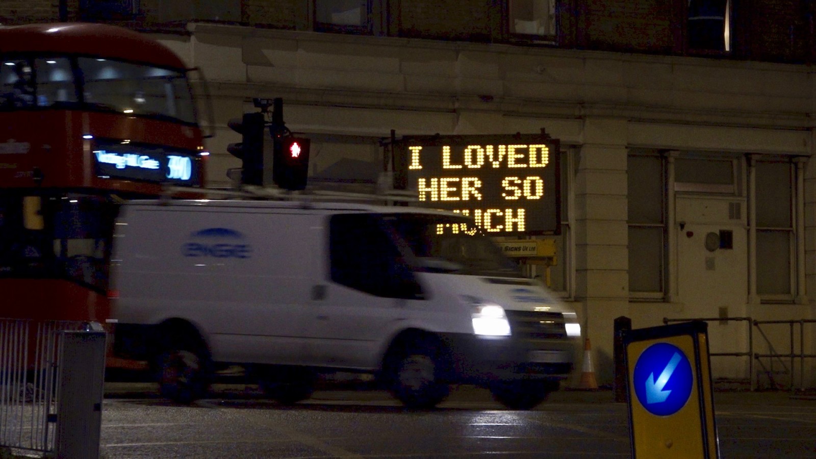 A white van drives in front of an illuminated traffic sign which reads 'I loved her so much.' The van is followed by a red double decker bus and the image has been taken at nighttime.