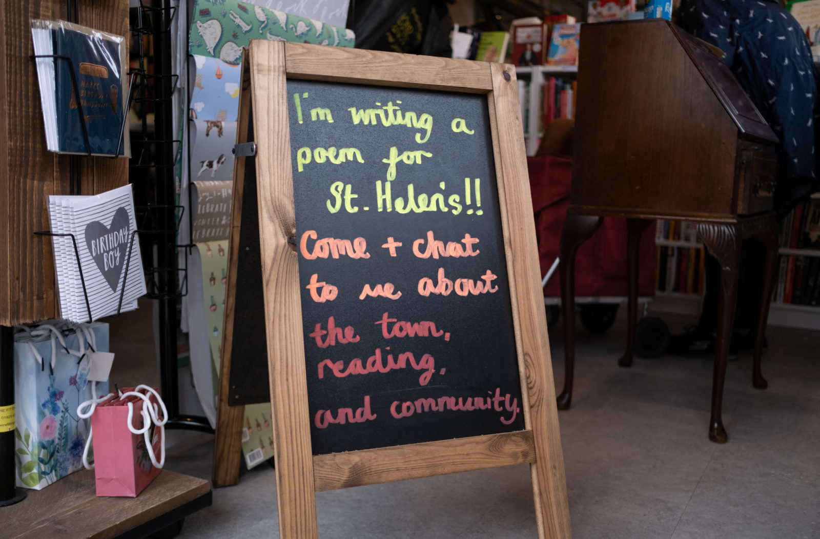 A board sign with writing on it saying "I'm writing a poem for St Helens!! Come + chat to me about the town, reading and community."