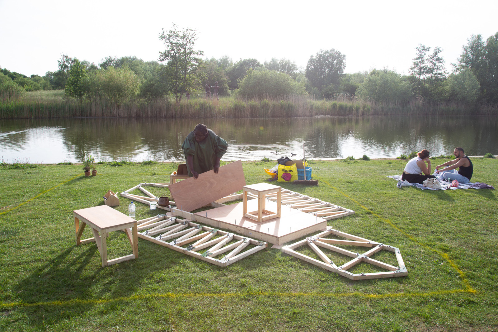Sonia Hughes building a small wooden structure on the grass in front of a lake.