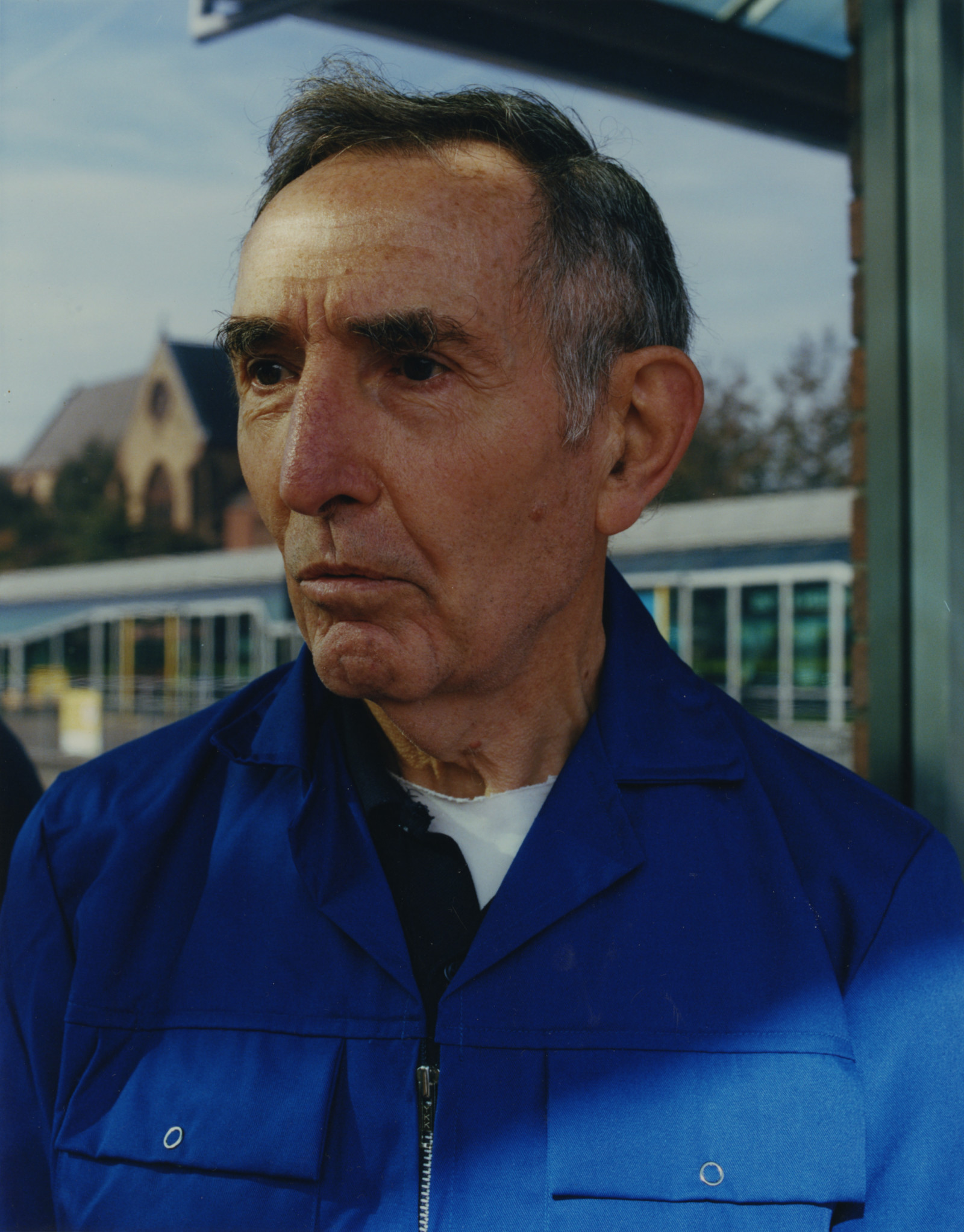 A middle aged man with dark hair, wearing a blue jacket stands at a bus stop