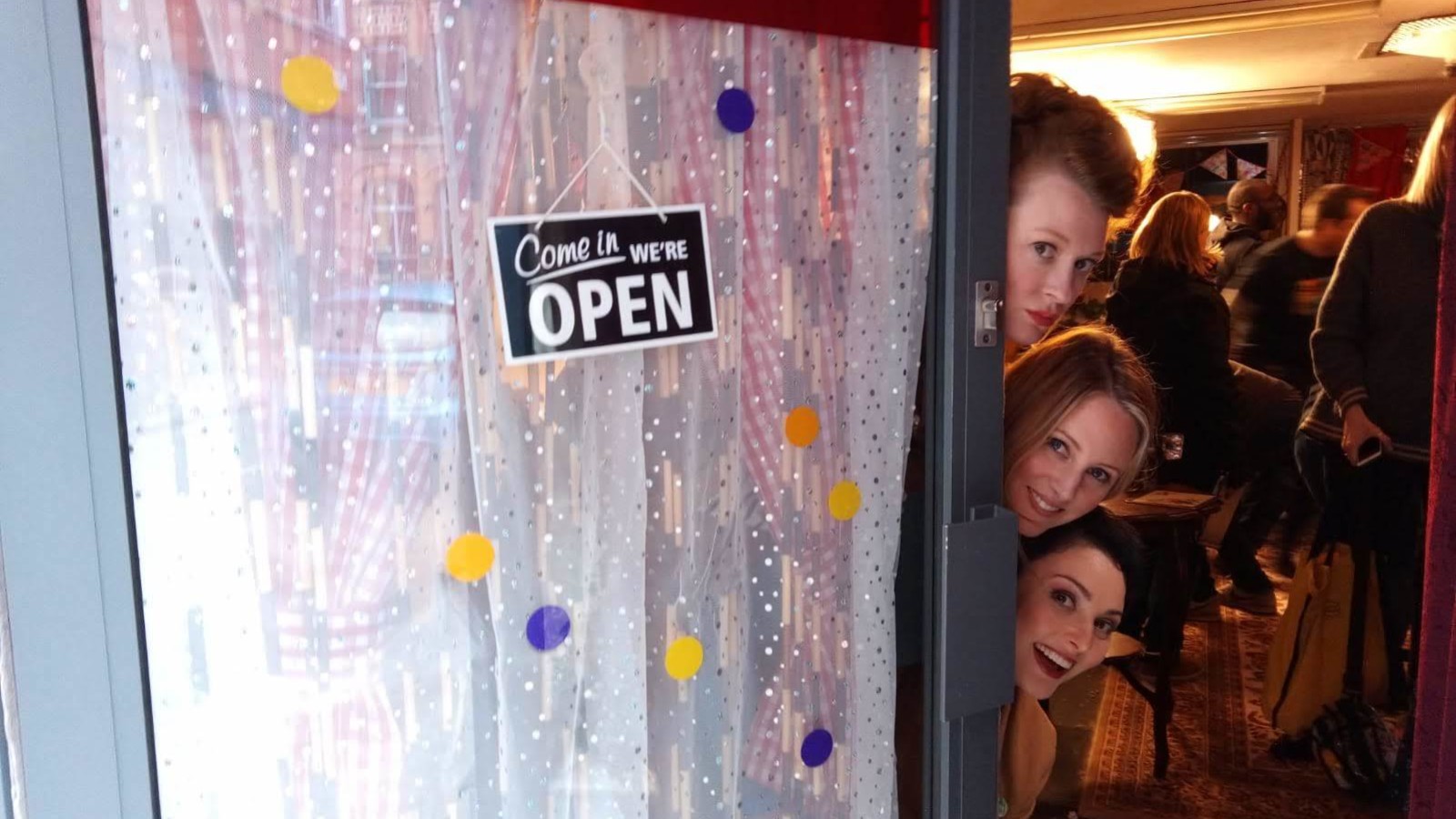 The artists peering around a glass door with the sign "come in, we're open" on it.