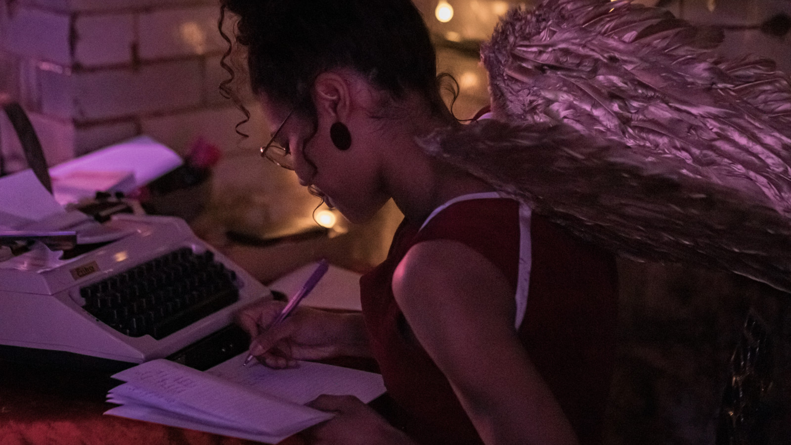 A young woman is sat at a table writing in a notepad. Next to her is a typewriter. She is wearing silvery angel wings.