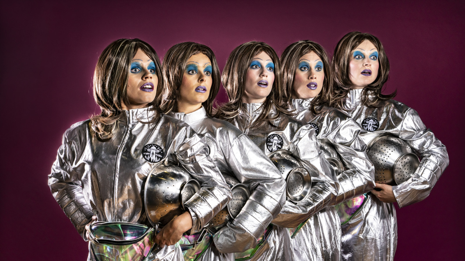 Five people stand in a row. They are wearing shiny silver suits and have bold blue eyeshadow.