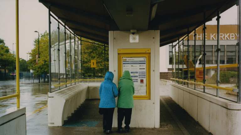 Two people stand with their backs turned away from the camera at a bus station in Kirkby