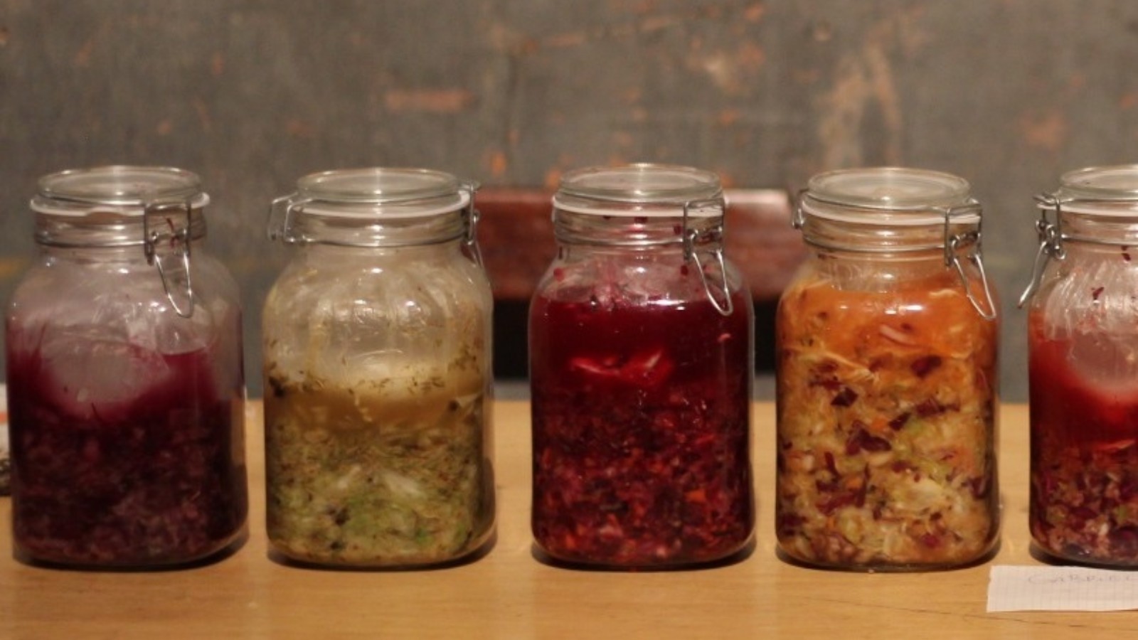 A line of jars are on a wooden table, each has different fermented foods in it.