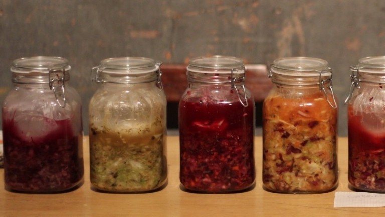A line of jars are on a wooden table, each has different fermented foods in it.