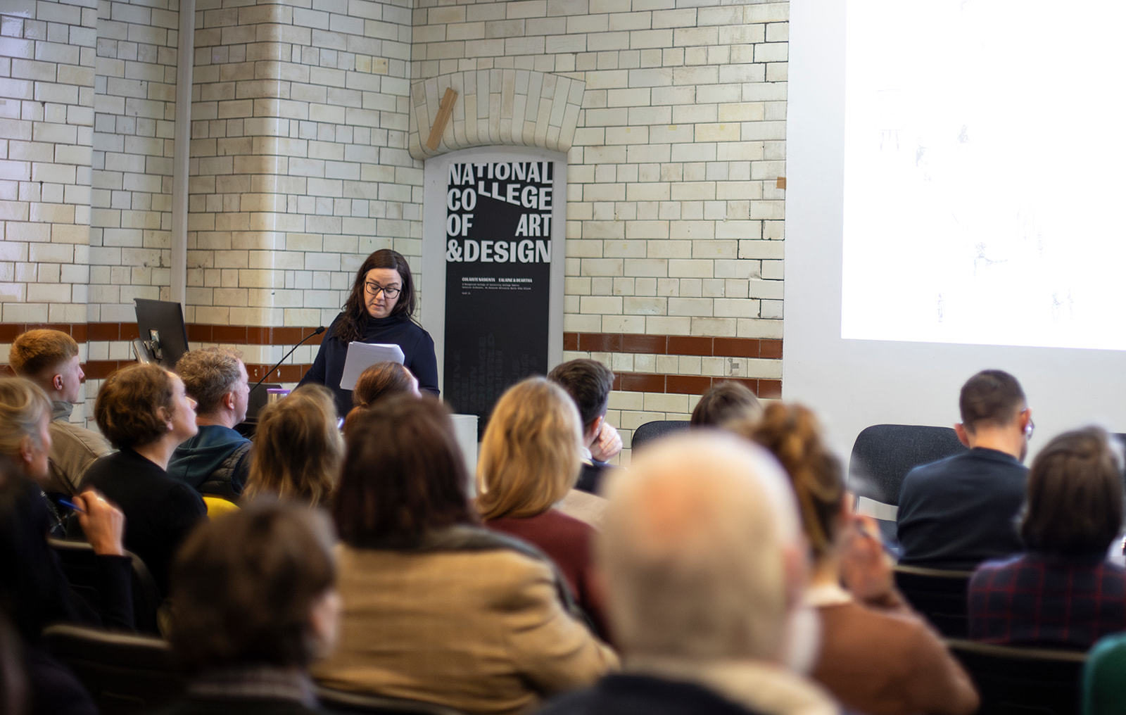 A woman with dark hair and a black jumper stands reading from a paper in front of an audience