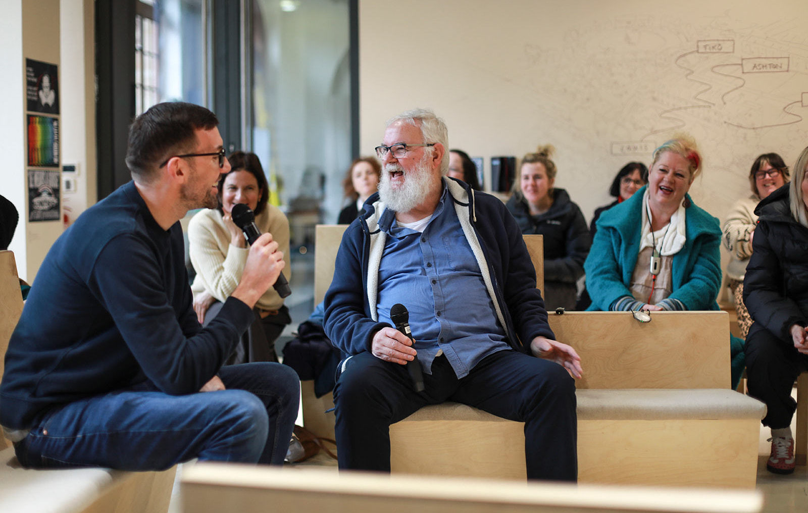 Two men, n with dark brown hair and one with white hair and beard sit in conversation in a naturally lit room. They are holding microphones and laughing. In the background a number of people watch on laughing