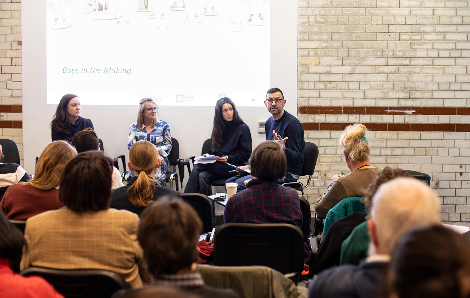 Four people sit in chairs on a stage in front of a big screen that reads 'Boys in the Making', there is an audience watching.
