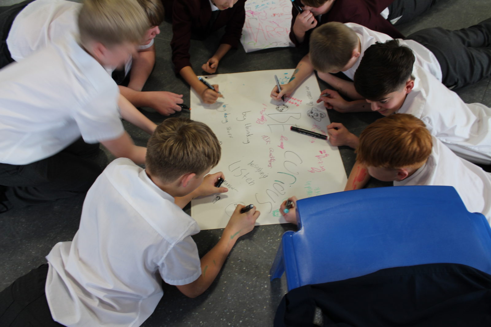 A group of teenage boys in school shirt and trouser lie the ground huddled around a large piece of paper. They are writing on the paper, which has the words 'be curious' and other writing on it.