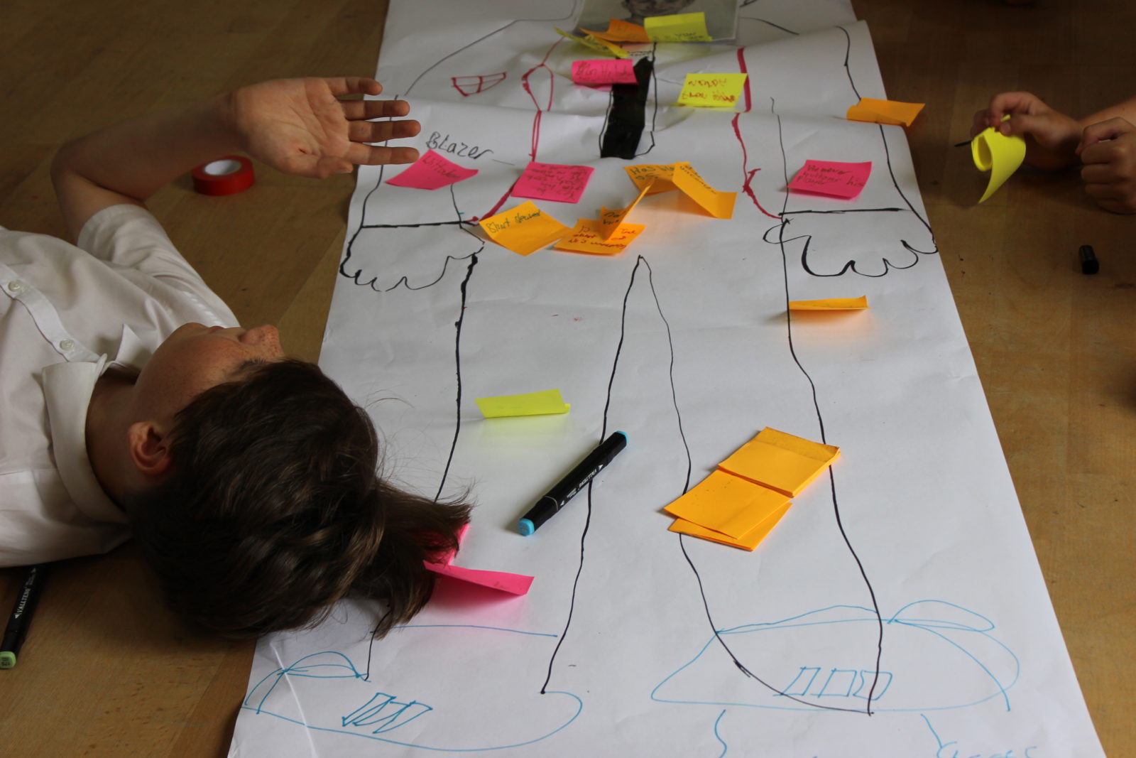 A teenage boy in school uniform lies on his back on the floor. Under his head is a piece of paper with a life size outline in marker pen, with coloured sticky notes scattered around the outline.