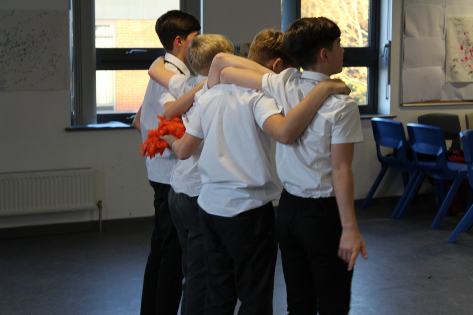 A row of four boys stand in a line with their arms around each others shoulders. We see the back of their heads as they look towards something off camera.