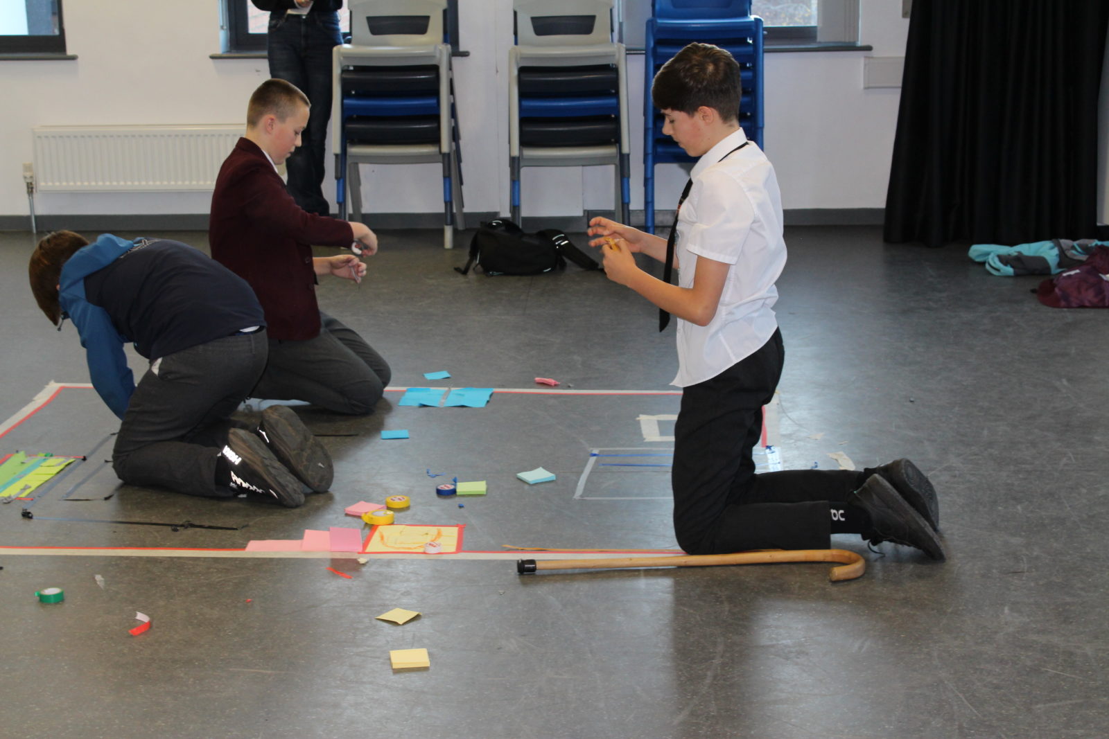 Three teenage boys in school uniform kneel on a vinyl floor. There is an arrangement of coloured tape and paper making a large shape on the floor and they are adding to the arrnagement.