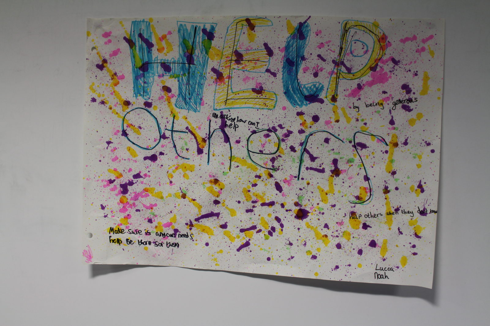 A large sheet of paper on a wall with colourful speckles and splatters of paint. Writing on the paper reads 'Help Others' in large font, with smaller similar phrases dotted around it.