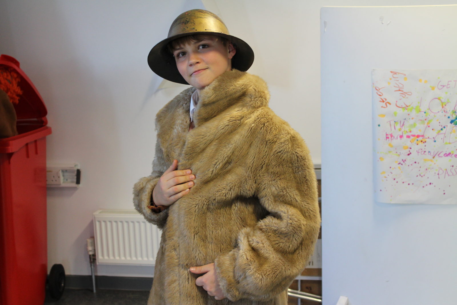 A teenage boy stands looking at the camera. He wears a light brown fur coat and a metal (gold) helmet.