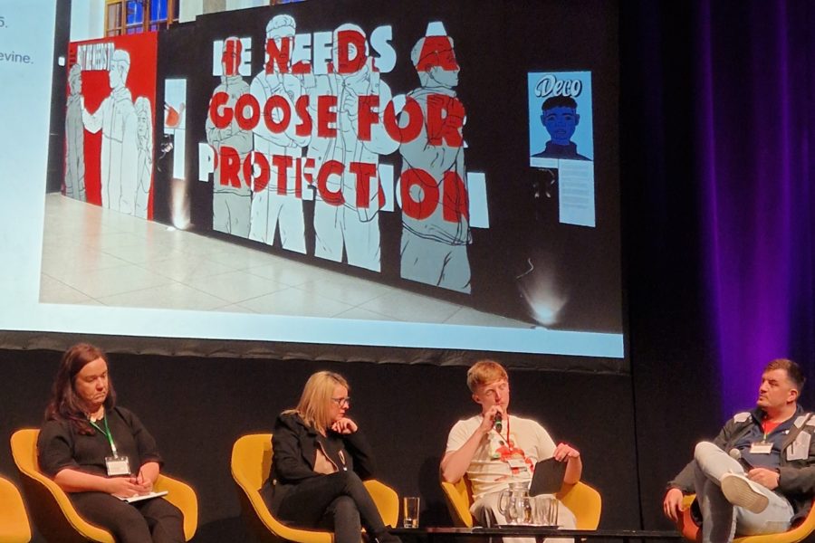 Four people sit on a stage around a coffee table with water glasses. Behind them is a large screen showing a photograph of an exhibition wall.