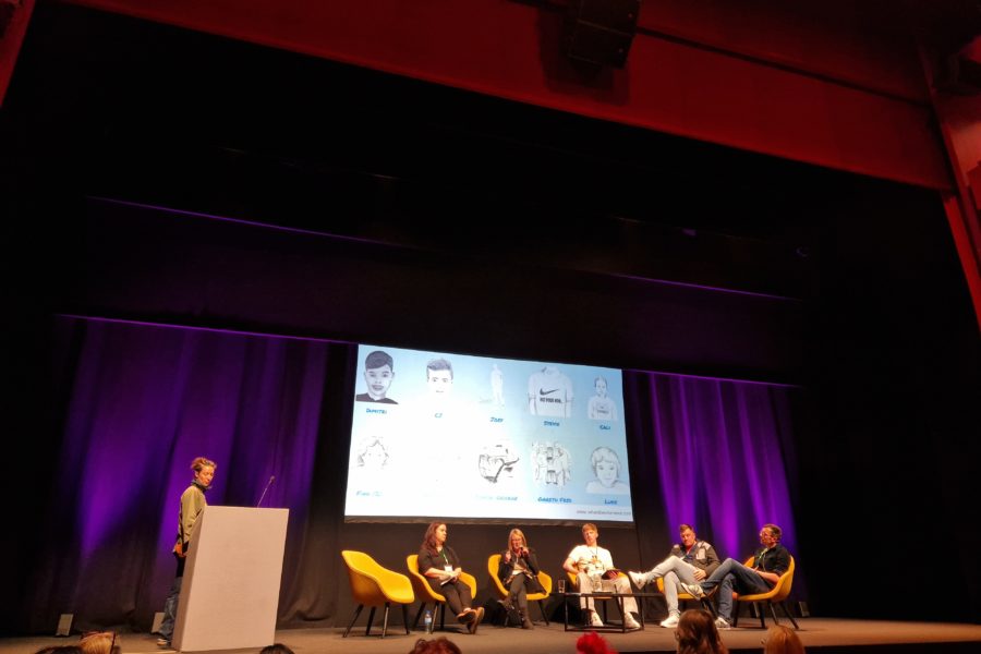 Five people sit on a stage around a coffee table with water glasses. Behind them is a large screen showing a 10 pencil drawings of boys with a name under each boy.