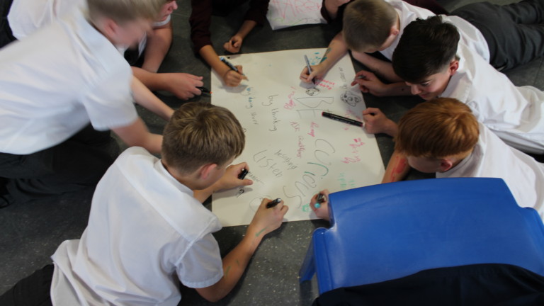 A group of teenage boys in school shirt and trouser lie the ground huddled around a large piece of paper. They are writing on the paper, which has the words 'be curious' and other writing on it.