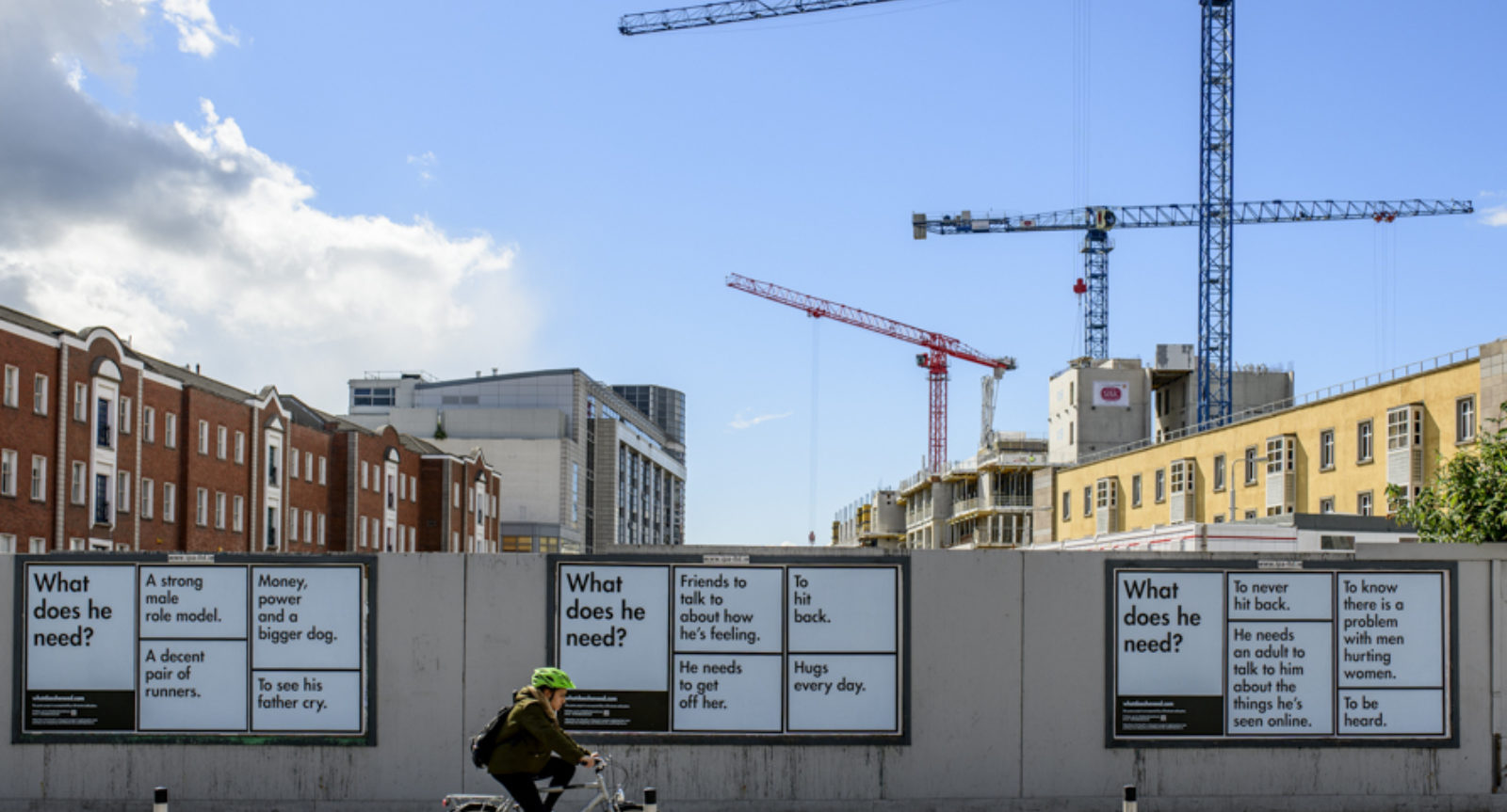 A series of black and white text based posters on an  outdoor wall asking "what does he need?". Cranes in the background and a cyclist passes in front