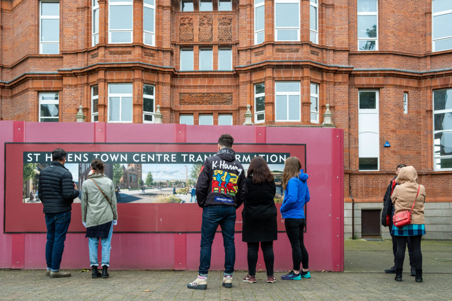 Five people stand with their back to us looking at a large, temporary display in front of a large red brick building. The display, partly obscured, reads St Helens Town Centre Transformation