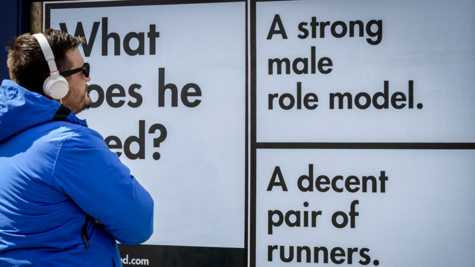 A series of black and white text based posters on an  outdoor wall asking "what does he need?". A man in a blue jacket wearing sunglasses and headphones reads the posters.