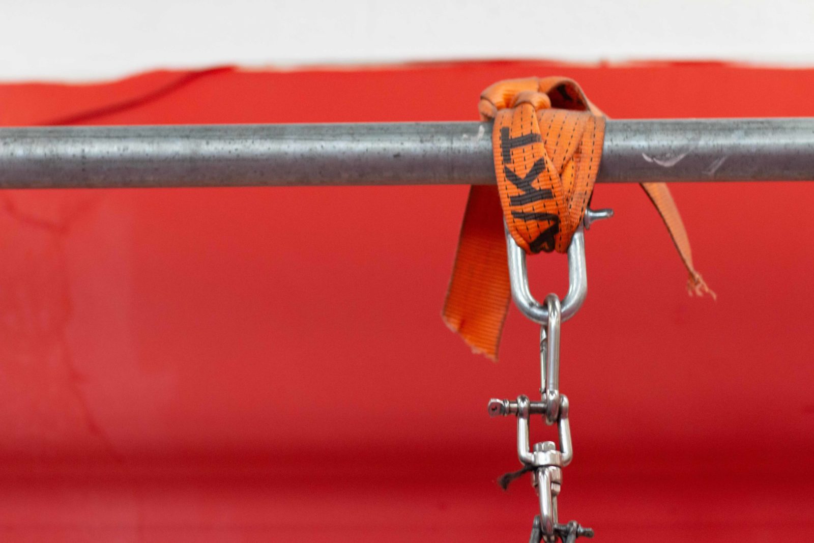 A metal bar against a red and white background. An orange strap is wrapped around the bar with a carabiner attached.