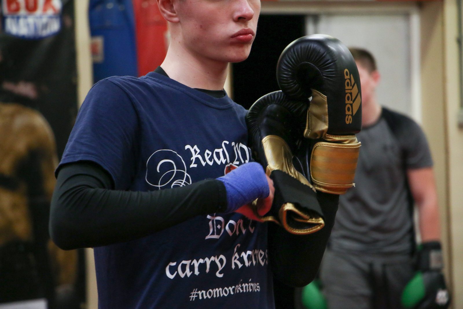 A person wearing a navy t-shirt and black long sleeved top pulls on a pair of black and gold boxing gloves.