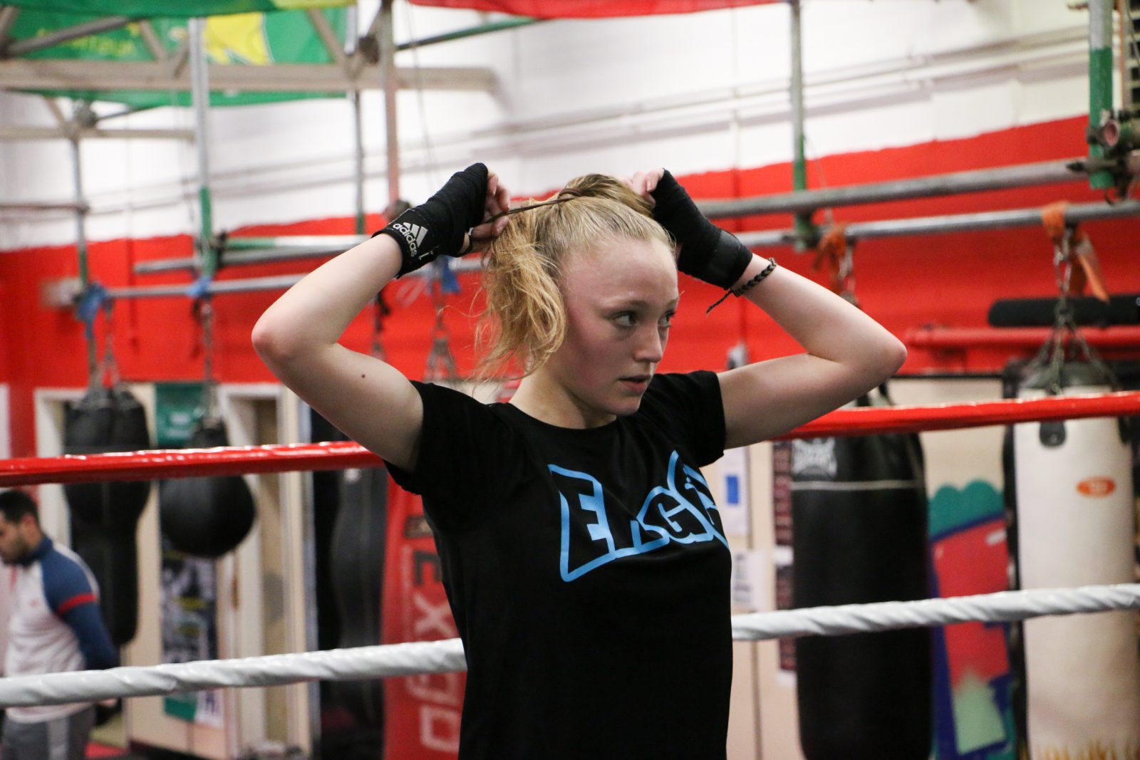 A young person with blonde hair wears a black t-shirt and thing black, fingerless gloves. They are tying their hair in a ponytail.