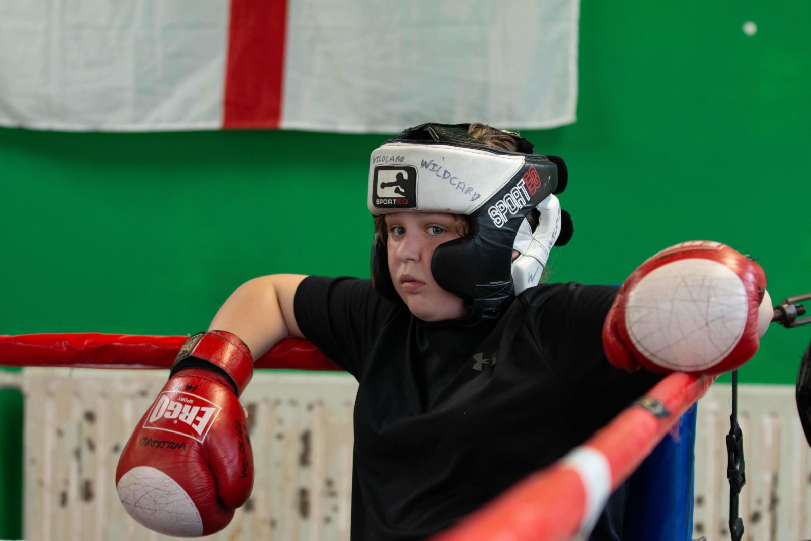 A young person leans against the inside corner of a boxing ring. They are wearing a black and white headguards and red and white boxing gloves.