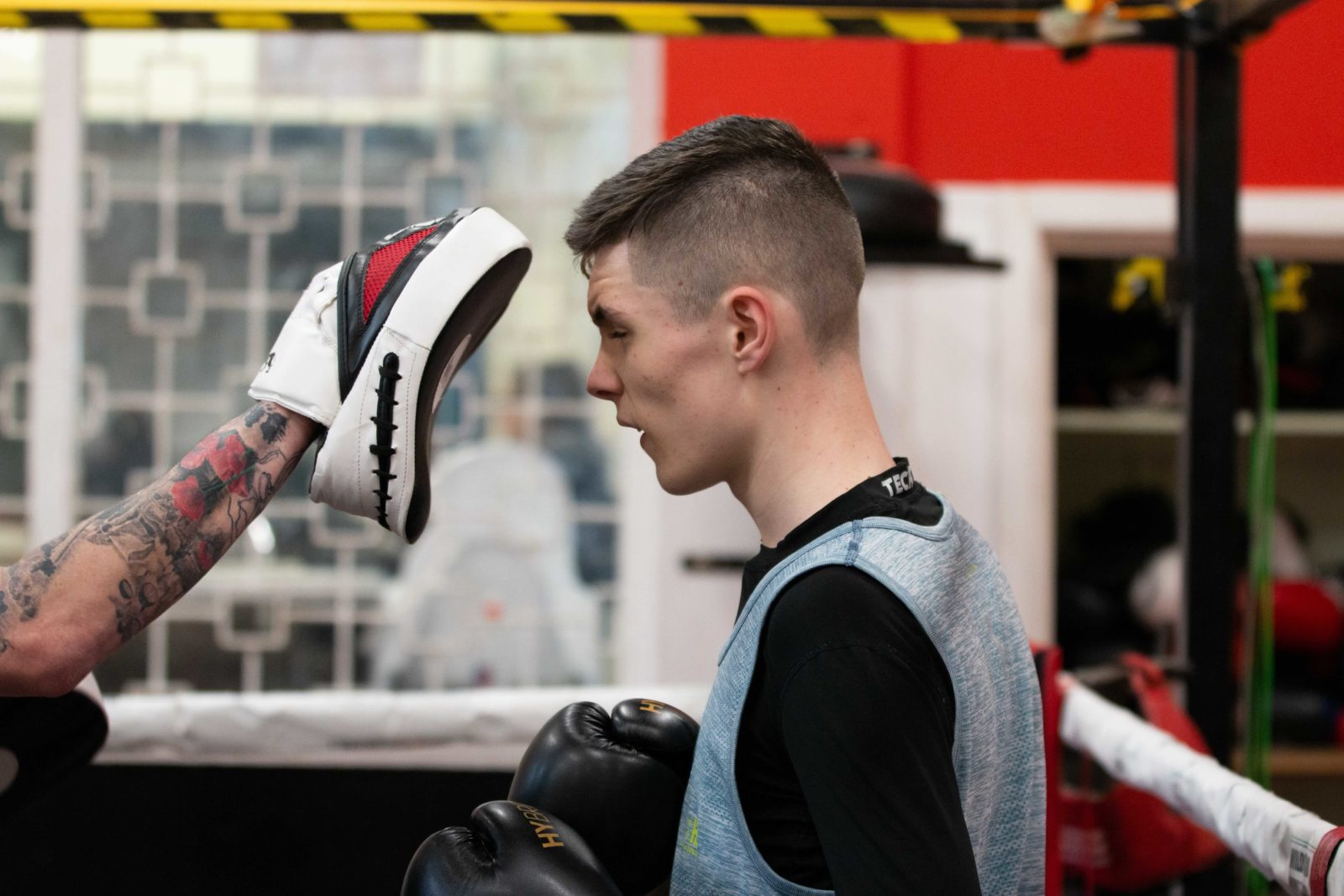 A young person wears a black long sleeve top, a blue sports vest and black boxing gloves. A tattooed arm holds a boxing pad close to their face.
