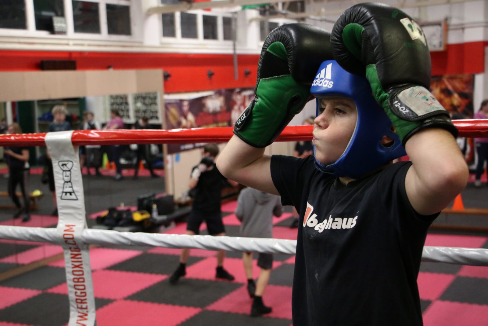 A young person wearing a blue boxing headguard and black and green boxing gloves holds their hands on top of their head in a boxing ring. In the background two people are boxing on a black and red chequered floor.